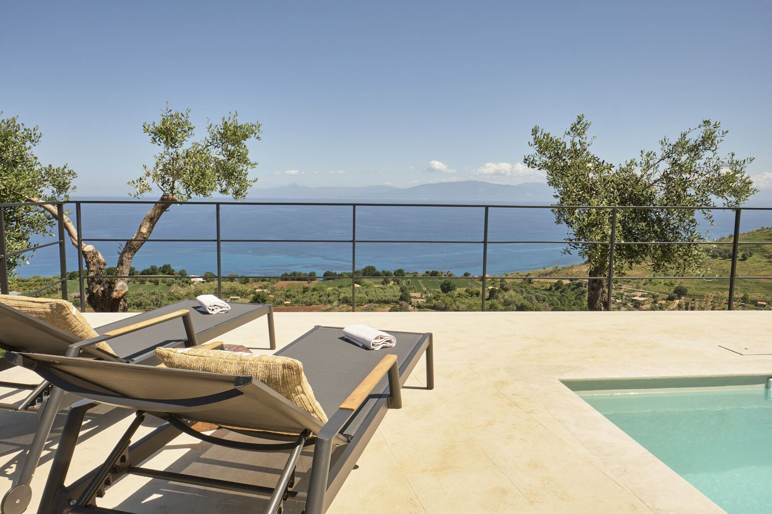 Luxury outdoor seating area with sun loungers overlooking a scenic ocean view and lush landscape, featuring a private pool and olive trees. Perfect for relaxation and enjoying nature. | Only on SopranoVillas