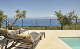 Luxury outdoor seating area with sun loungers overlooking a scenic ocean view and lush landscape, featuring a private pool and olive trees. Perfect for relaxation and enjoying nature.