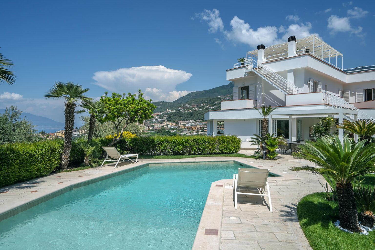 Luxurious villa with a swimming pool surrounded by lush greenery and palm trees, set against a scenic mountain backdrop. Ideal for relaxation and outdoor living. | Only on SopranoVillas
