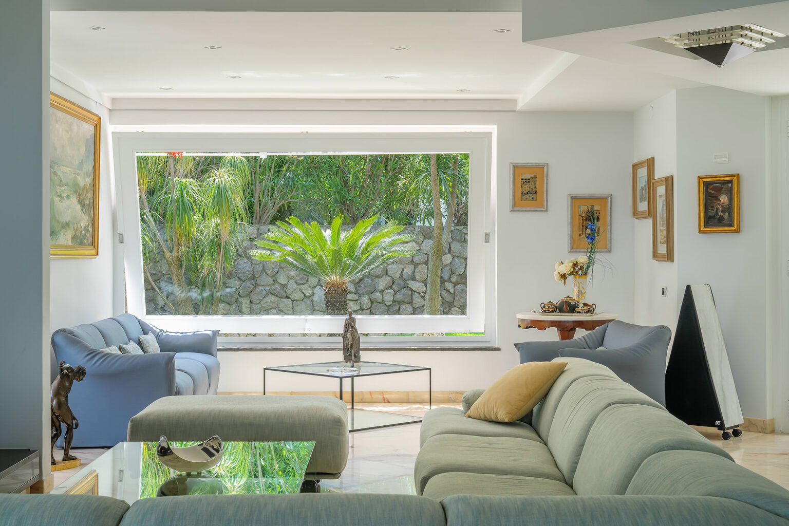 Spacious modern living room featuring large windows with views of a lush garden, comfortable blue sofas, and stylish decor. The room is adorned with framed artworks and a coffee table, creating a bright and inviting atmosphere. | Only on SopranoVillas