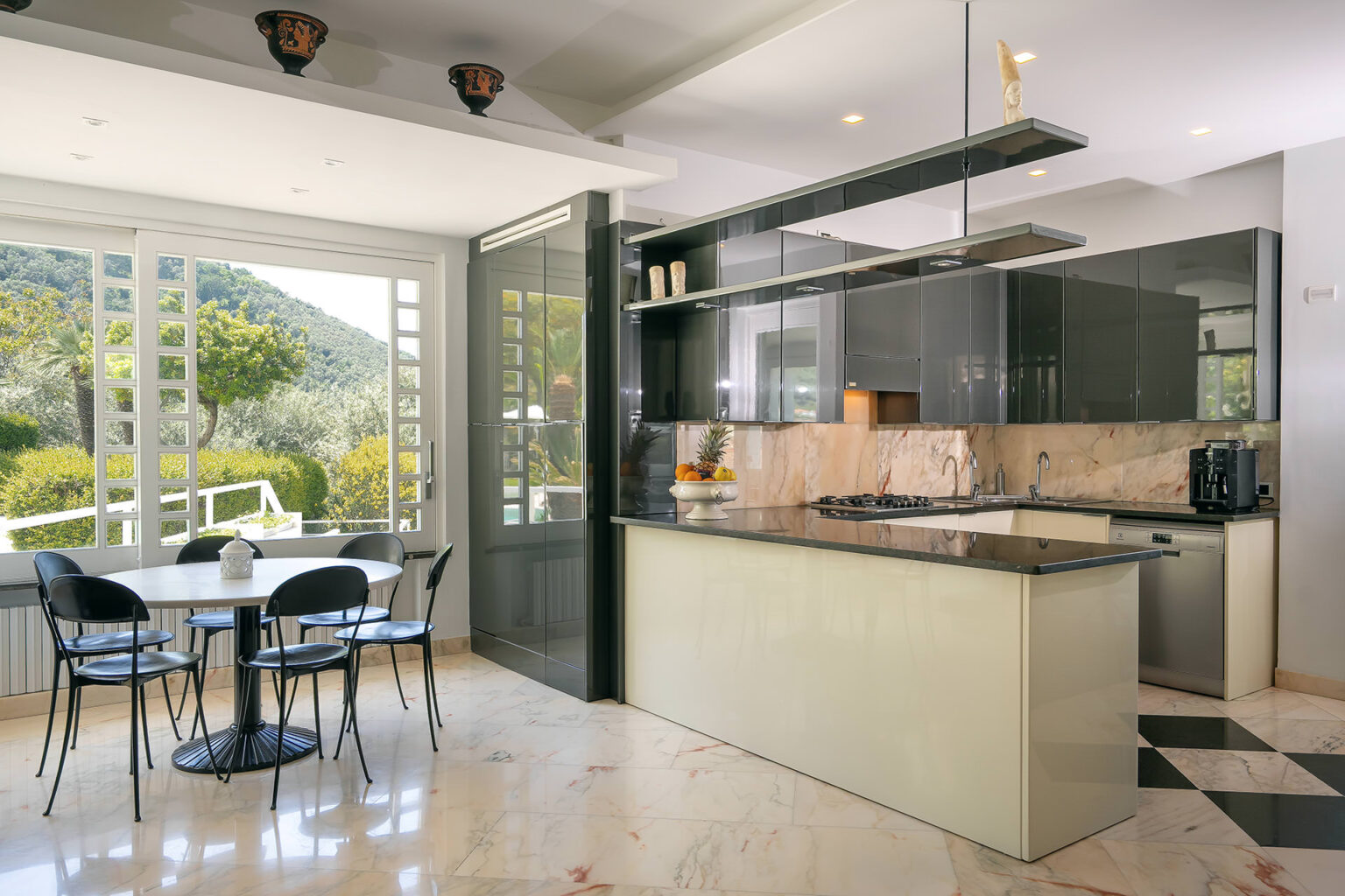 Modern kitchen with a sleek design featuring a marble countertop, black cabinetry, and an open layout connecting to a dining area with a round table and chairs. Large windows provide natural light and views of the outdoors. | Only on SopranoVillas