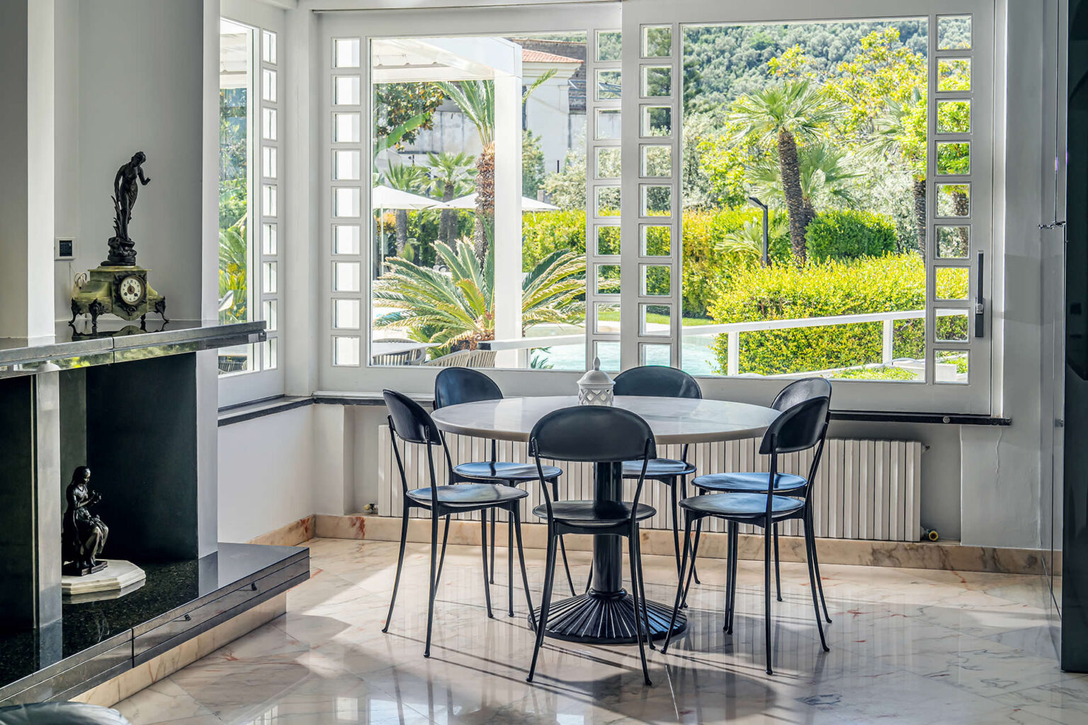Bright and airy dining area featuring a round table surrounded by black chairs, with large windows showcasing a lush outdoor garden and palm trees. Elegant decor includes a decorative clock and a statue on the fireplace, complemented by marble flooring. Ideal for modern interior design inspiration. | Only on SopranoVillas