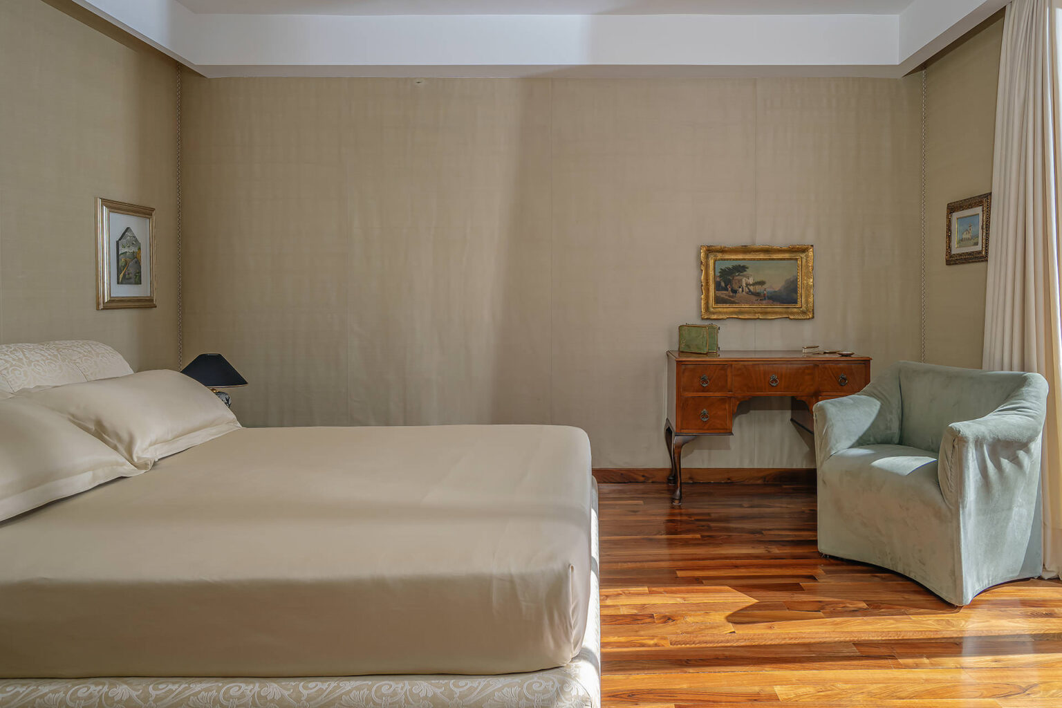 Cozy bedroom featuring a beige bedspread, a wooden side table, an armchair, and framed artwork on the walls, illuminated by natural light. | Only on SopranoVillas