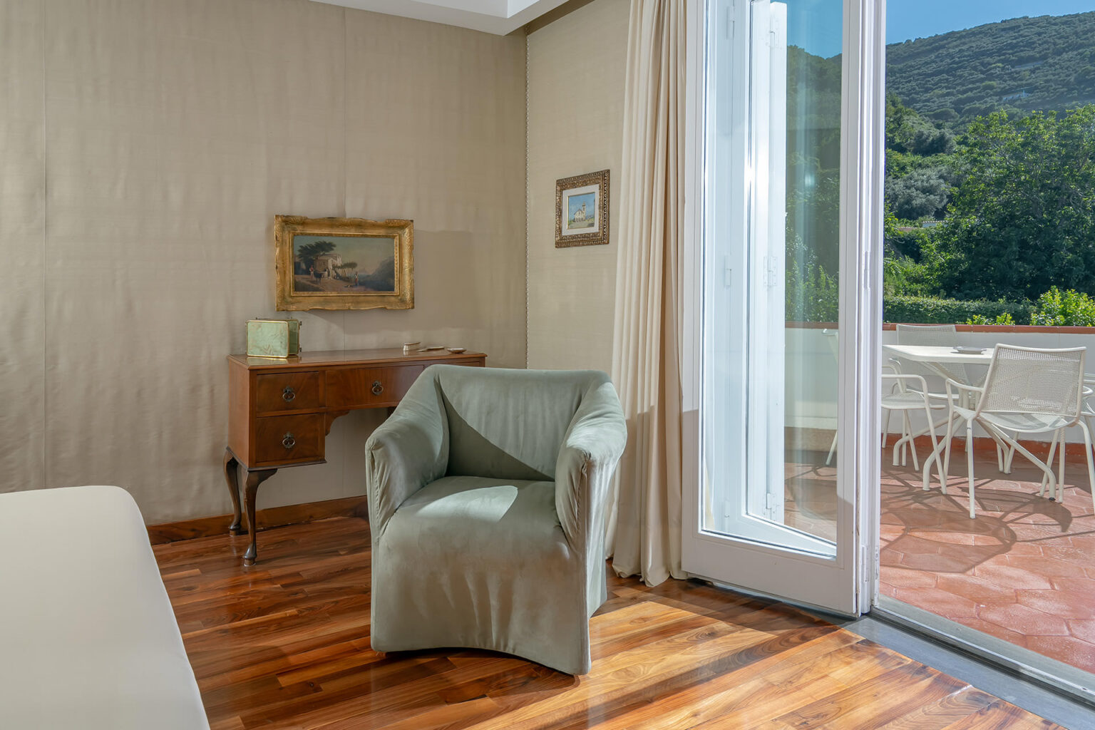 Cozy interior of a room featuring a green armchair, wooden flooring, and large glass doors opening to a scenic patio with outdoor seating. Decorated with a framed artwork, the space offers a warm and inviting atmosphere. | Only on SopranoVillas