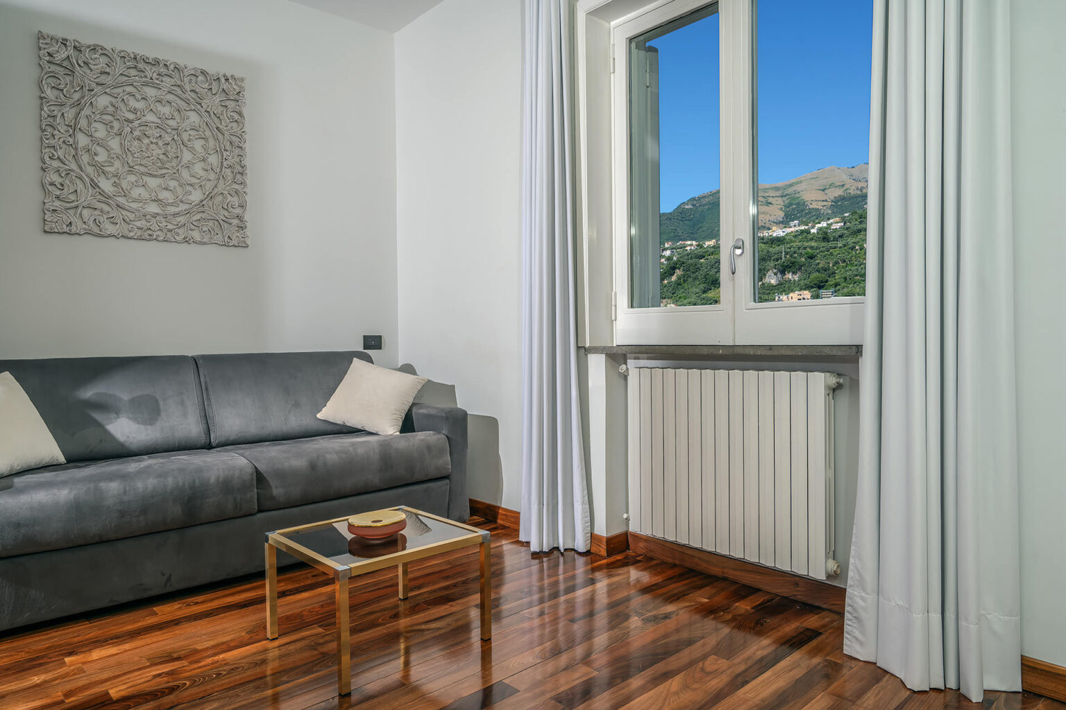 Modern living room with a gray sofa and decorative pillows, featuring a stylish gold coffee table. Large window offers a scenic view of mountains, complemented by white curtains and polished wooden flooring. Ideal for cozy relaxation and natural light. | Only on SopranoVillas