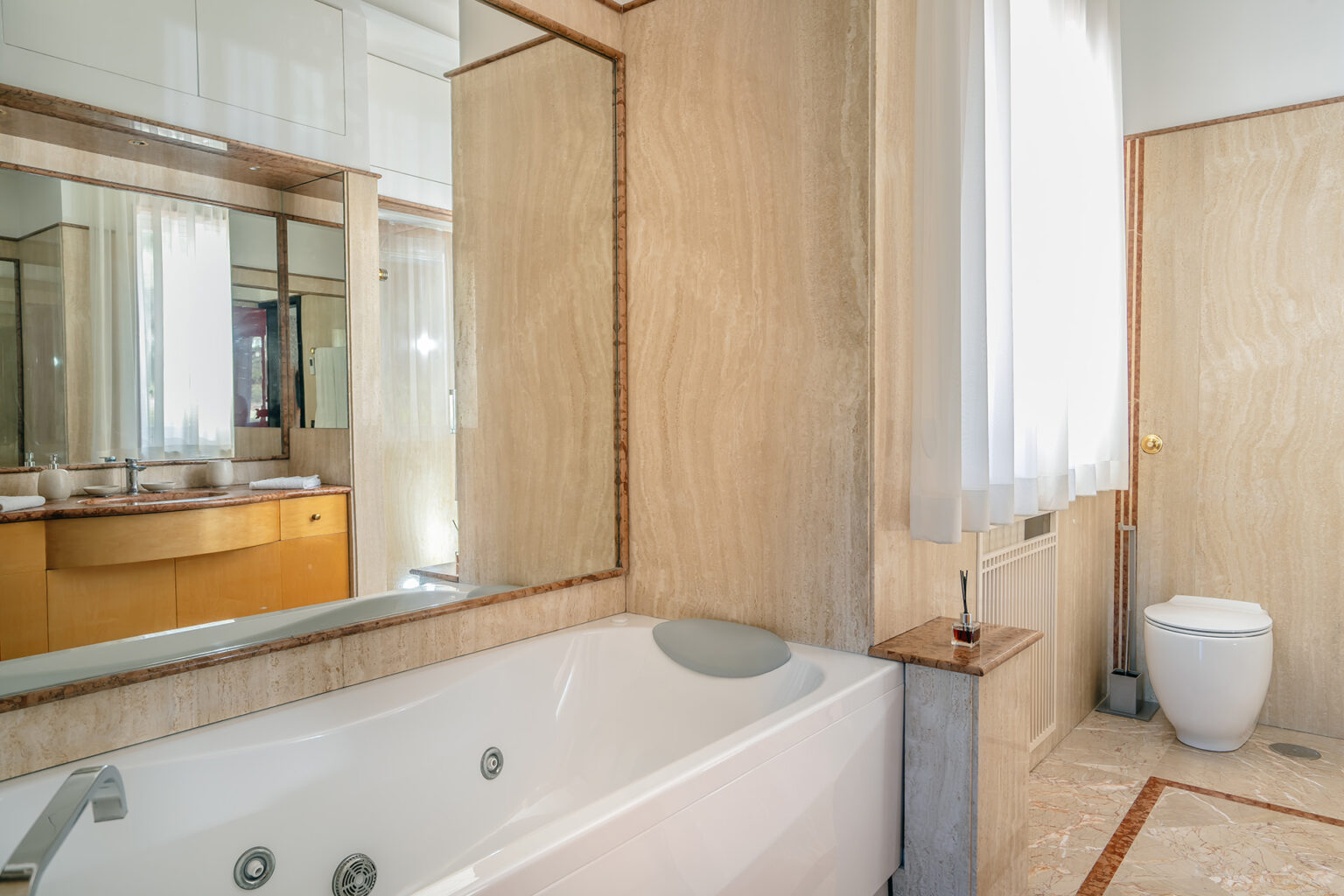 Modern bathroom interior featuring a luxurious bathtub, large mirror, and elegant wooden cabinetry, complemented by natural light from a window. The design emphasizes clean lines and a contemporary aesthetic. | Only on SopranoVillas