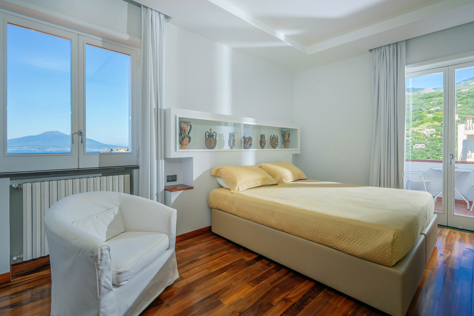 Modern bedroom with a yellow bedspread, white walls, and wooden flooring, featuring a cozy armchair and large windows showcasing a scenic view of mountains and greenery. | Only on SopranoVillas