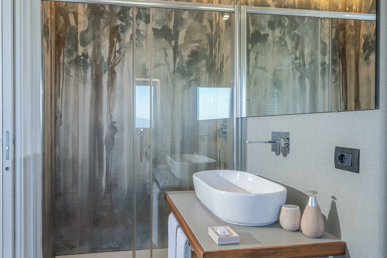 Modern bathroom featuring a sleek glass shower, a stylish white vessel sink on a wooden countertop, and contemporary decor elements. Natural light enhances the serene ambiance, complemented by a large wall mirror. | Only on SopranoVillas