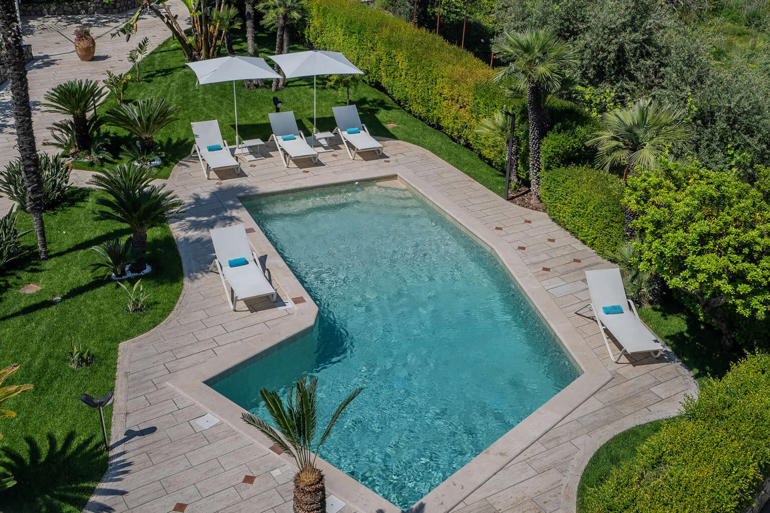 Aerial view of a serene outdoor swimming pool surrounded by lush greenery, featuring white lounge chairs and sun umbrellas for relaxation. | Only on SopranoVillas