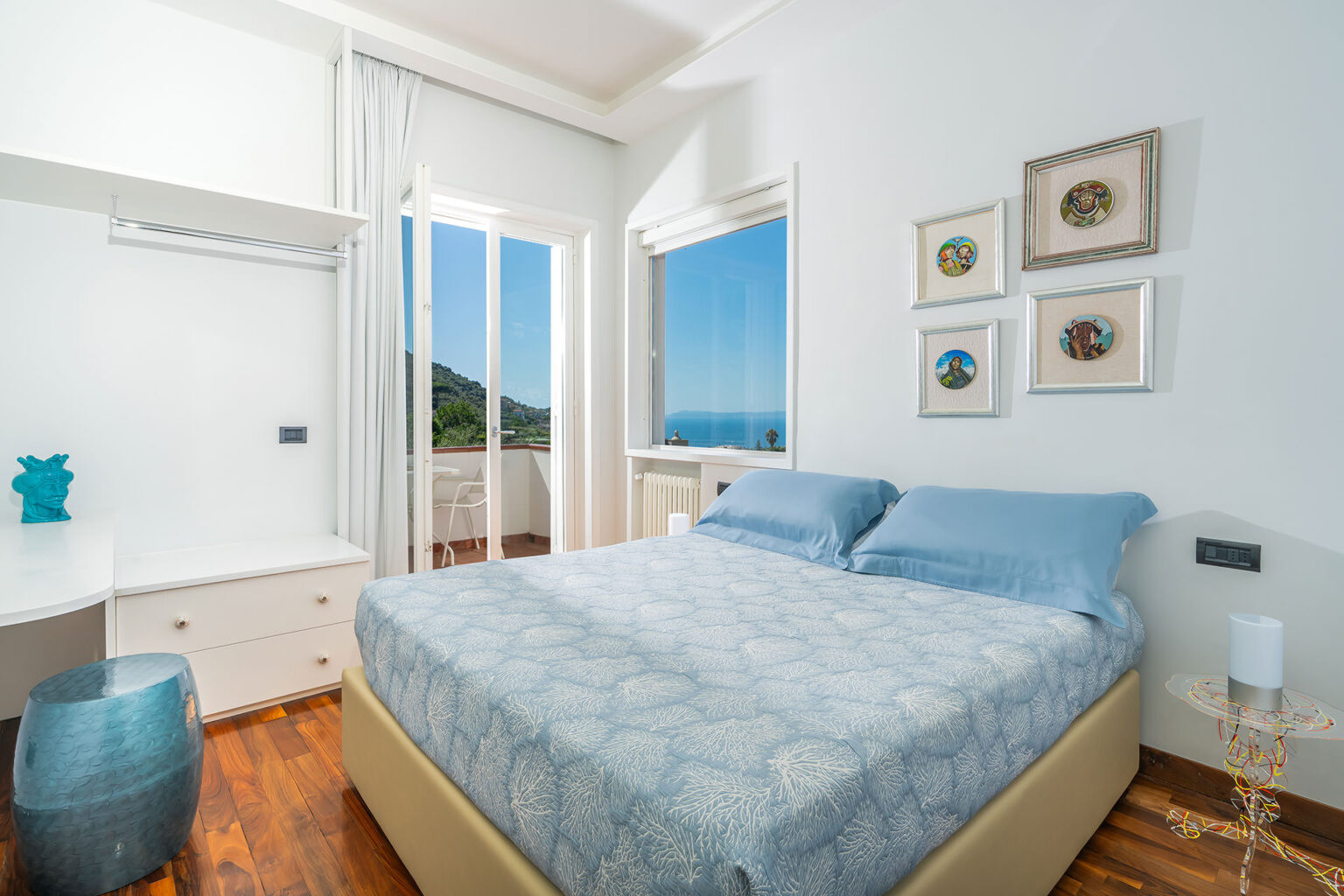 Cozy bedroom with a blue-themed decor, featuring a comfortable bed, a stylish bedside table, and a window offering a scenic view of the outdoors. The room is bright and airy, with modern artwork on the walls and a minimalist design. | Only on SopranoVillas
