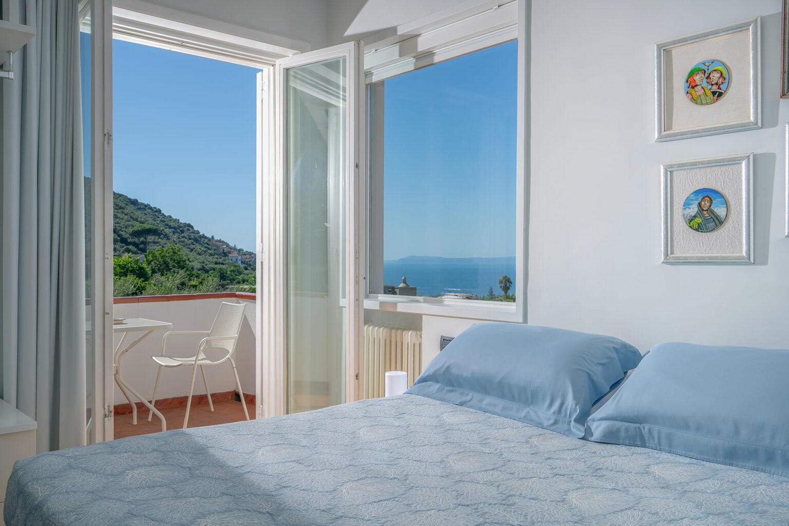 Cozy bedroom with blue bedding, featuring a large window that opens to a scenic view of the ocean and hills. A small balcony with outdoor seating is visible, enhancing the room's tranquil atmosphere. | Only on SopranoVillas