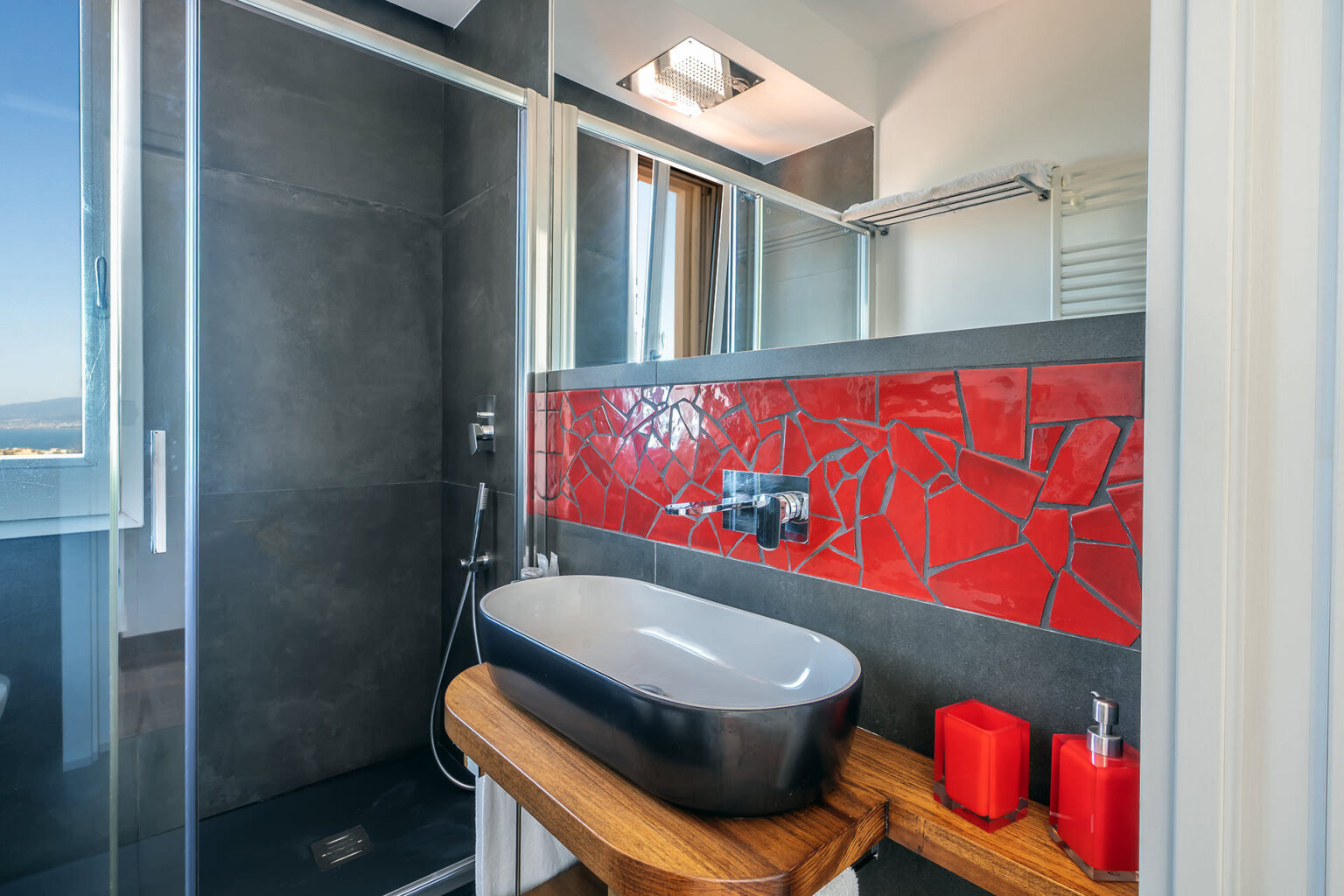 Modern bathroom featuring a sleek black freestanding sink, red mosaic accent wall, and glass shower enclosure, complemented by wooden shelving and stylish decor items. | Only on SopranoVillas