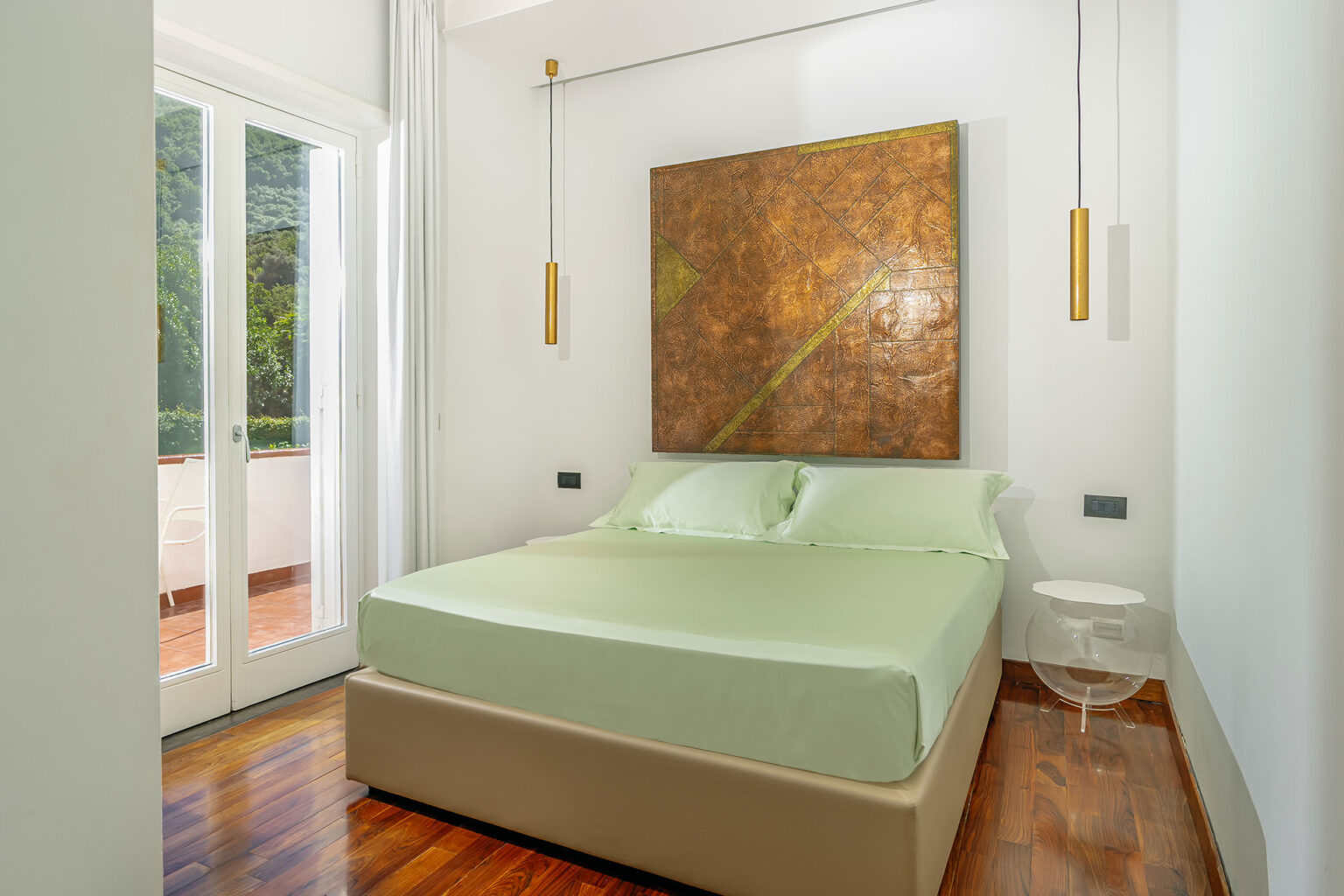 Modern bedroom featuring a light green bedspread, wooden flooring, and a large abstract painting on the wall. Natural light floods the space through sliding glass doors leading to a balcony. Elegant pendant lights add a stylish touch to the decor. | Only on SopranoVillas