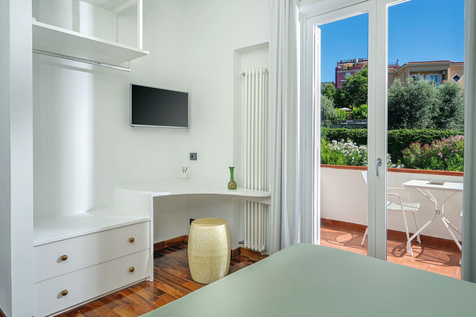 Modern hotel room featuring a minimalist design, equipped with a wall-mounted TV, a stylish desk area, and a cozy seating space. The room has large windows that open to a private balcony with views of lush greenery and a serene outdoor setting. The decor includes light-colored walls and wooden flooring, creating a bright and inviting atmosphere. | Only on SopranoVillas