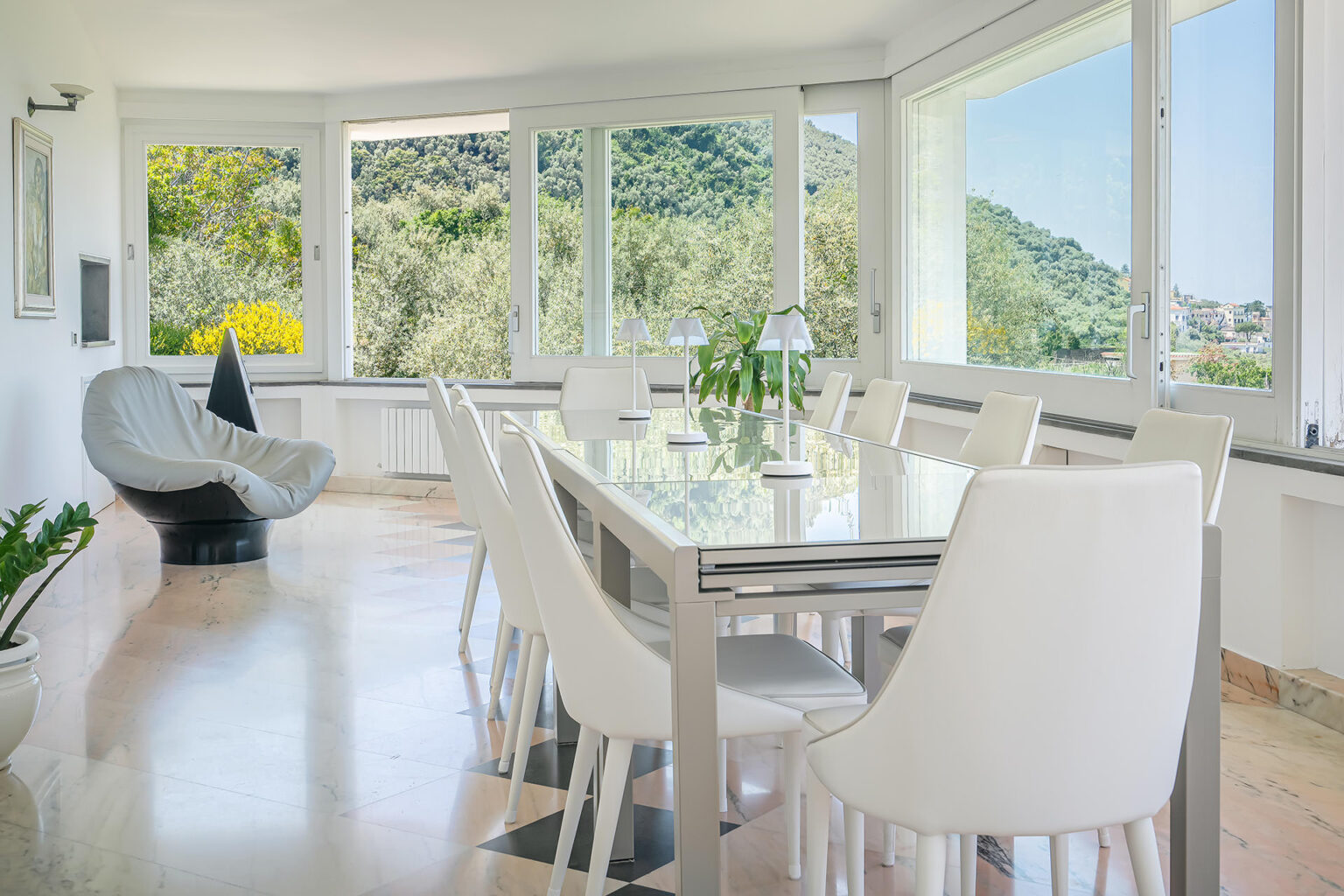 Modern dining room with a glass table and white chairs, featuring large windows that offer a scenic view of lush greenery and mountains. A stylish gray chair and potted plant enhance the contemporary decor, creating a bright and inviting atmosphere. | Only on SopranoVillas