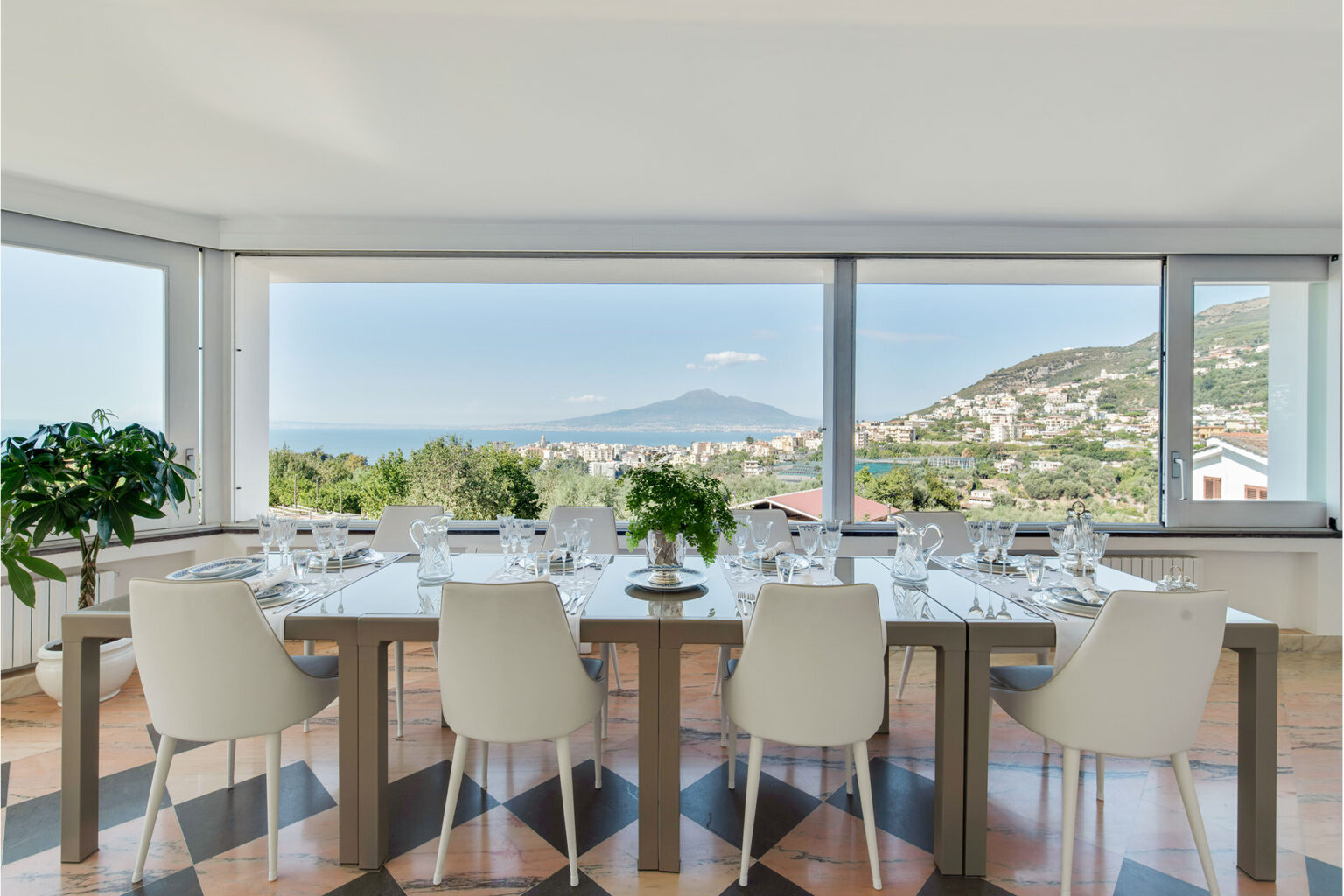 Modern dining room with a panoramic view of the coastline and Mount Vesuvius, featuring a sleek table set with elegant glassware and surrounded by white chairs, complemented by greenery and natural light. | Only on SopranoVillas