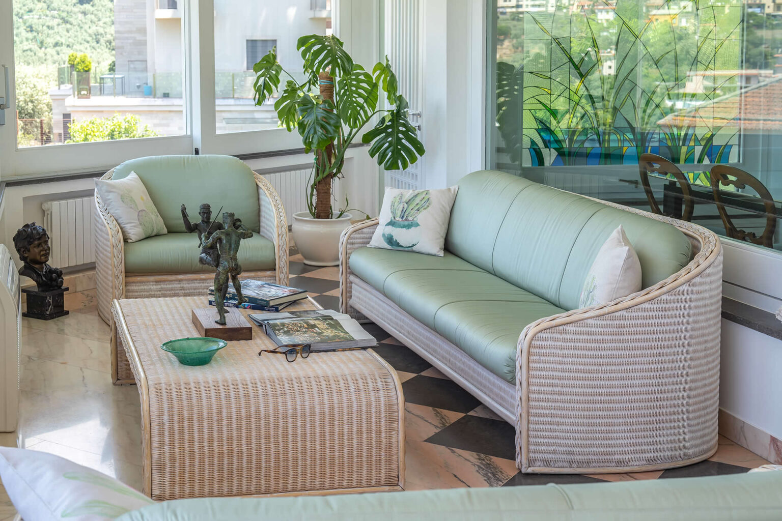 Bright and airy living room featuring a light green sofa and armchair, a woven coffee table, and decorative sculptures. The space is enhanced by large windows and a potted plant, creating a cozy and inviting atmosphere. | Only on SopranoVillas