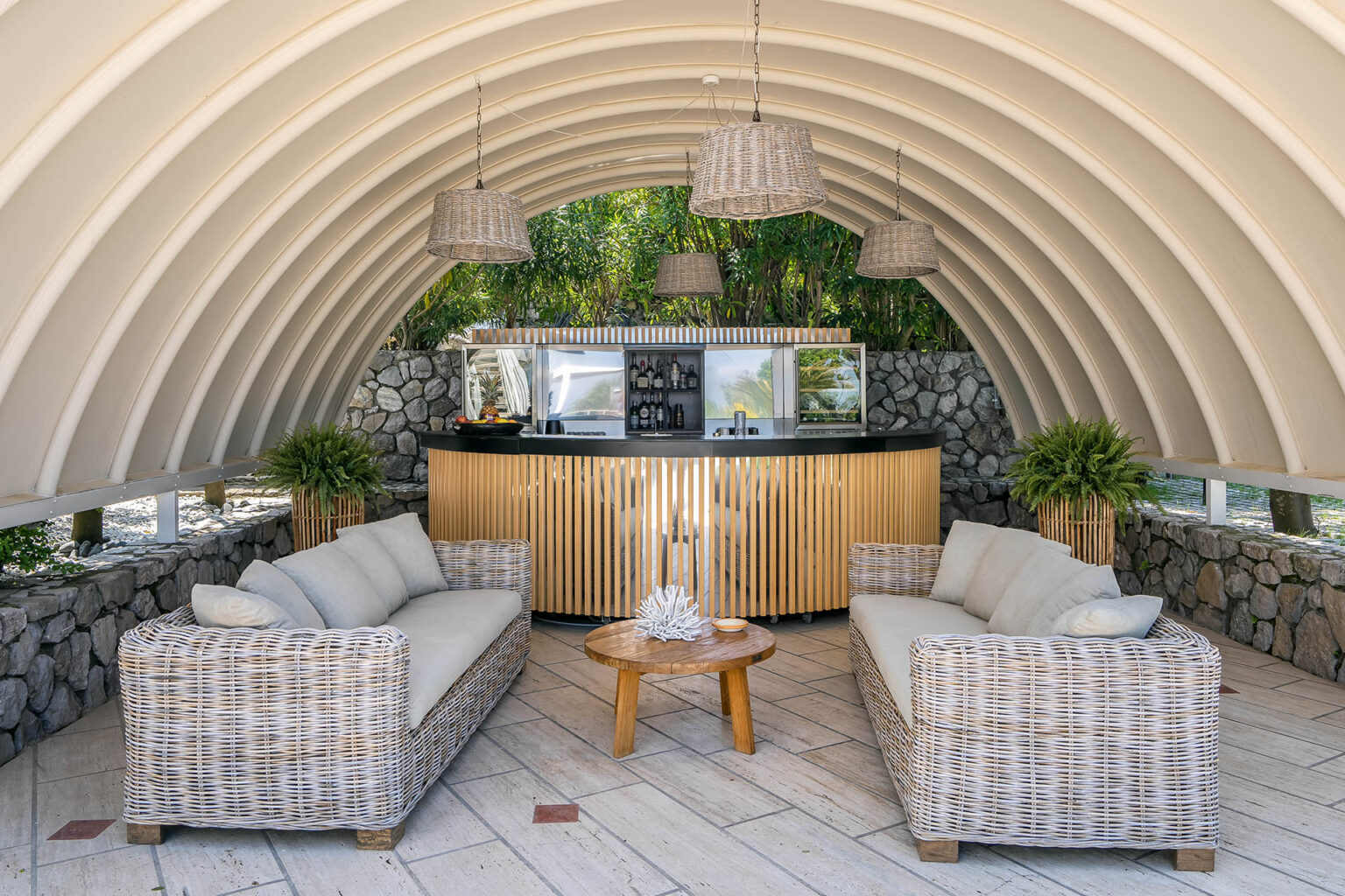 Cozy outdoor bar area featuring stylish wicker furniture, a wooden coffee table, and a modern bar counter with a stone backdrop, all under a curved, white canopy. Lush greenery enhances the tranquil atmosphere. | Only on SopranoVillas