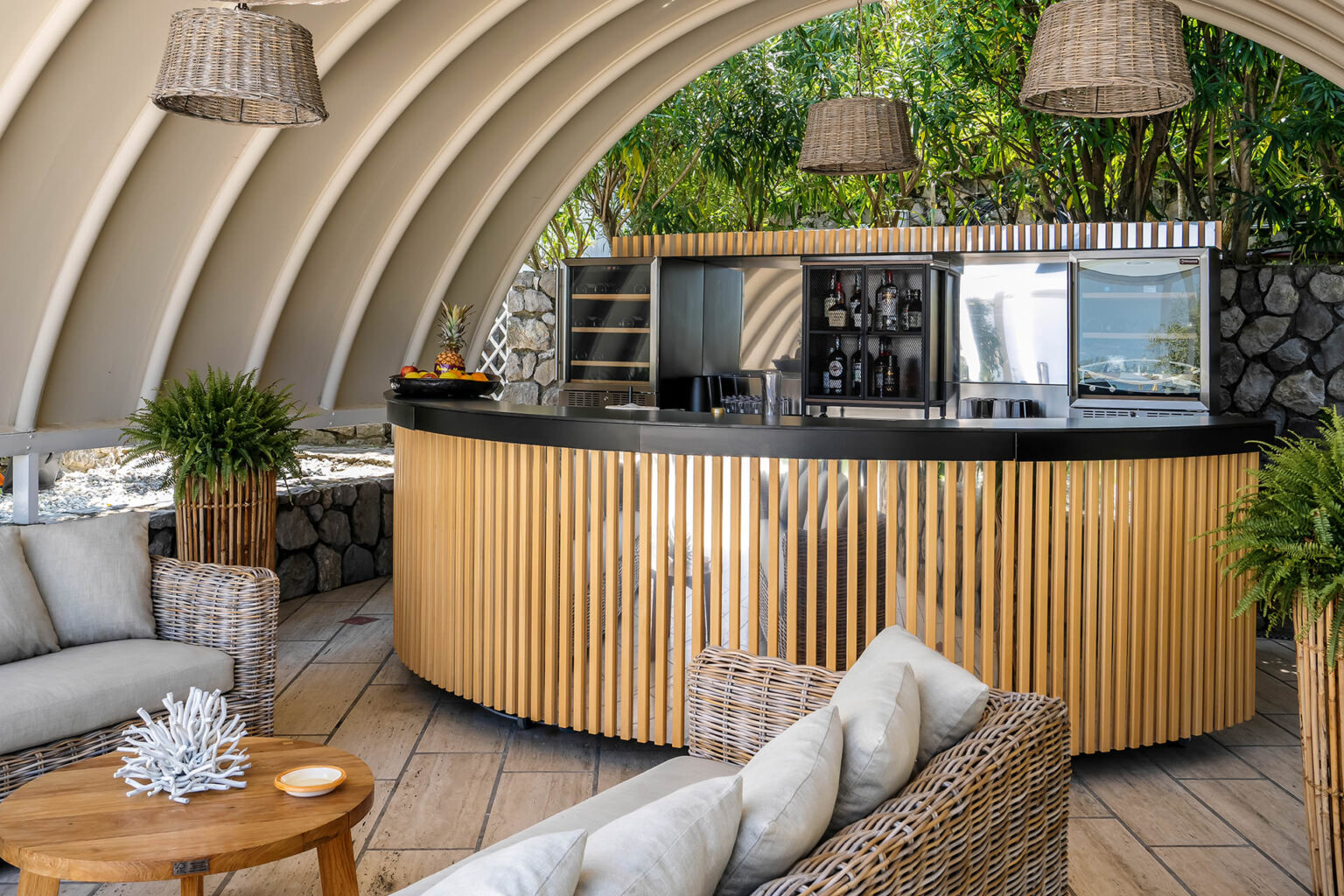 Stylish outdoor bar area featuring a curved wooden counter, modern appliances, and comfortable seating with wicker furniture, surrounded by lush greenery. Ideal for relaxation and social gatherings. | Only on SopranoVillas