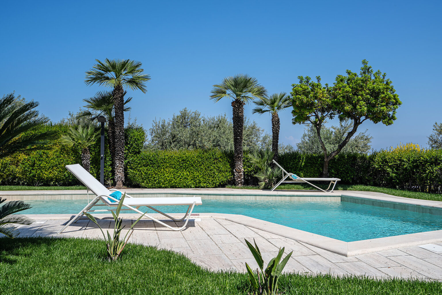 Serene outdoor swimming pool surrounded by lush greenery and palm trees, featuring comfortable lounge chairs under a clear blue sky. Perfect for relaxation and leisure activities in a tranquil setting. | Only on SopranoVillas