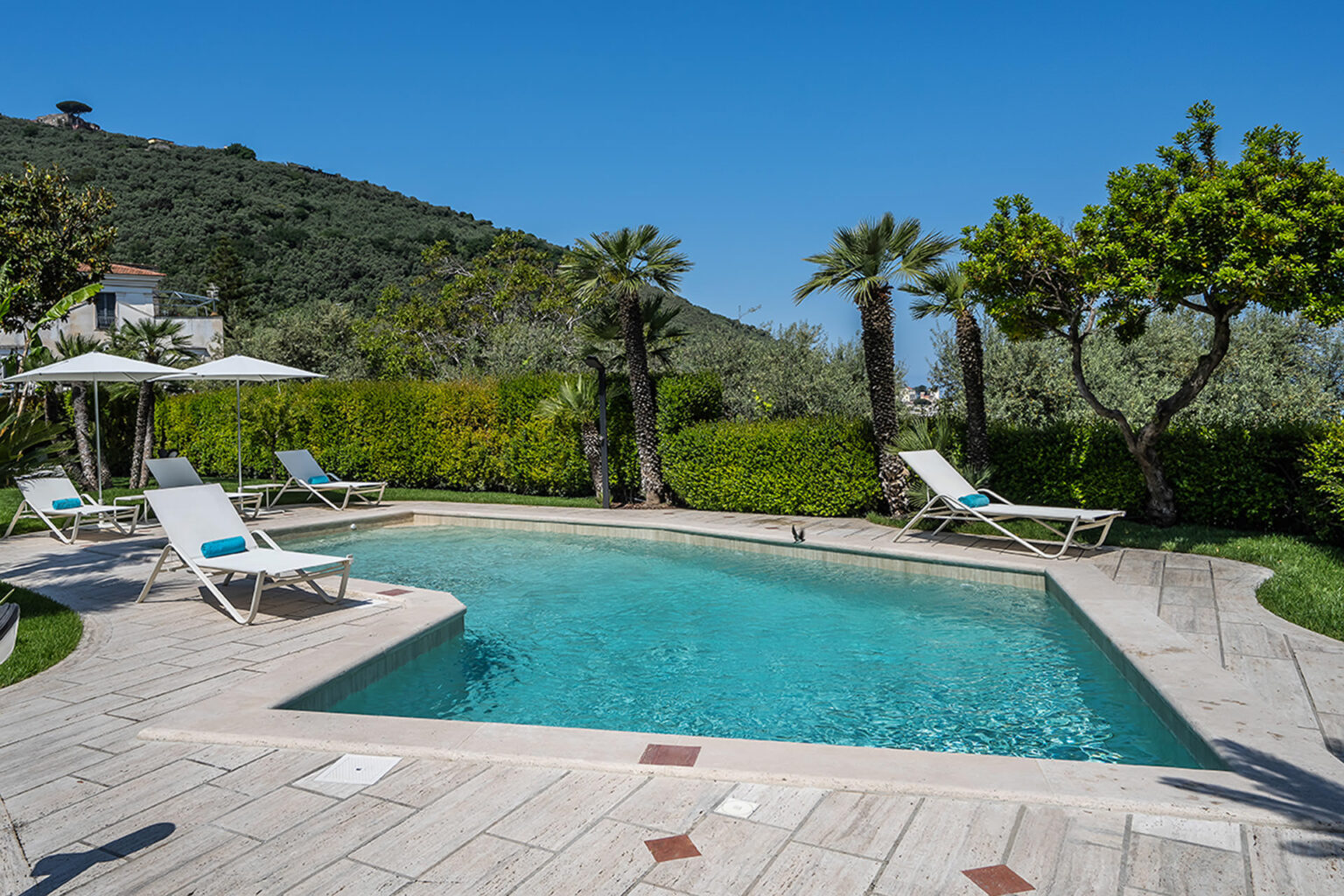 Luxurious outdoor pool area surrounded by lush greenery and palm trees, featuring sun loungers with blue cushions and umbrellas, perfect for relaxation in a serene setting. | Only on SopranoVillas