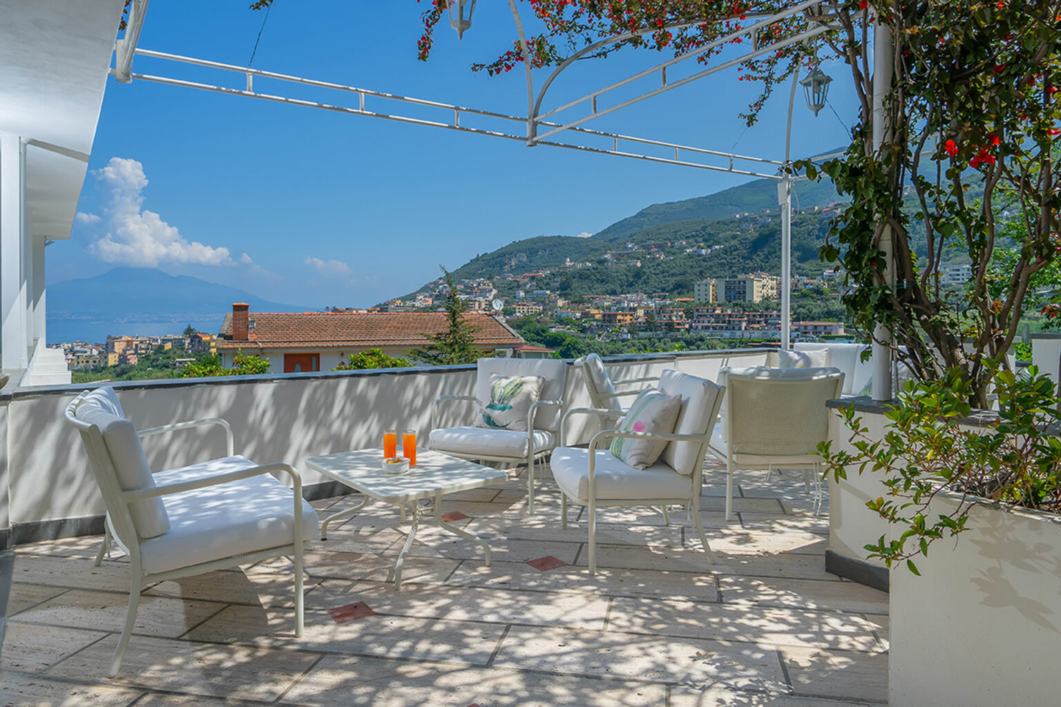Serene outdoor terrace with modern white furniture, overlooking a picturesque mountain view, perfect for relaxation and entertaining. Lush greenery and vibrant flowers enhance the tranquil ambiance under a clear blue sky. | Only on SopranoVillas