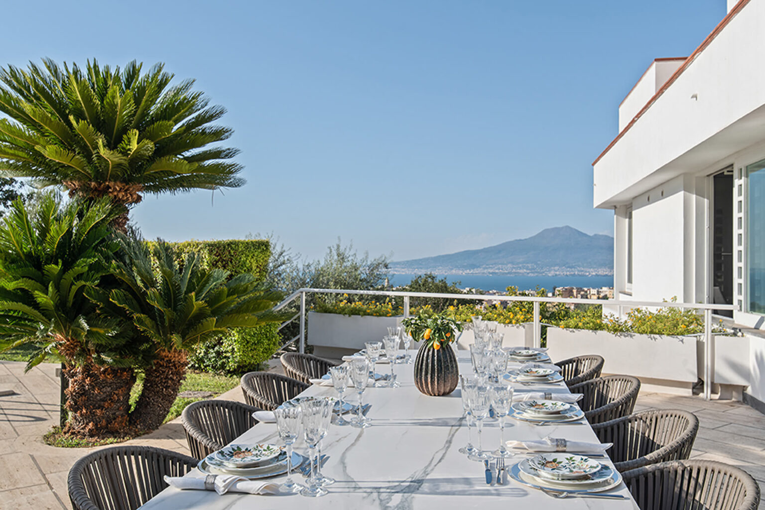 Outdoor dining setup with a long table and elegant tableware, surrounded by lush palm trees and a scenic view of the ocean and mountains in the background. Ideal for alfresco dining experiences. | Only on SopranoVillas