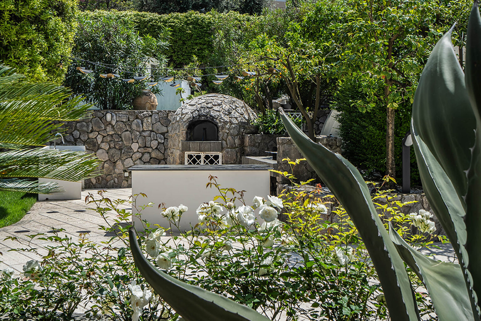 Lush garden featuring a stone pizza oven surrounded by greenery and blooming flowers, creating an inviting outdoor space for cooking and entertaining. | Only on SopranoVillas