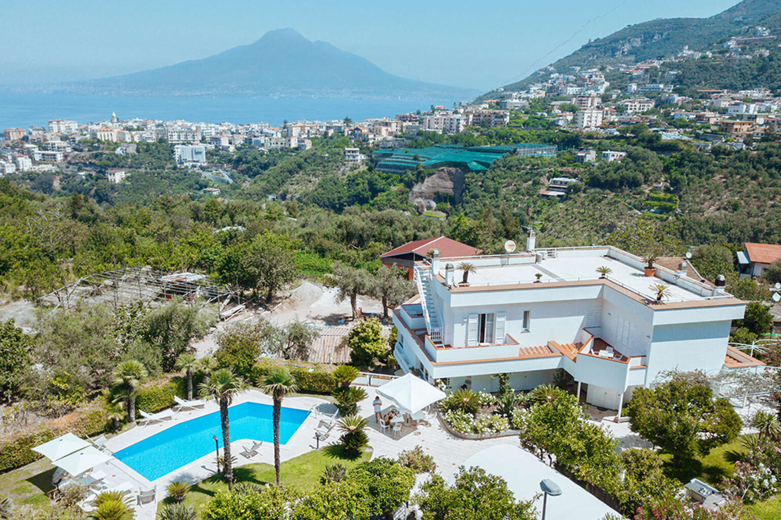 Aerial view of a modern villa with a swimming pool, surrounded by lush greenery and overlooking a coastal landscape with Mount Vesuvius in the background. | Only on SopranoVillas
