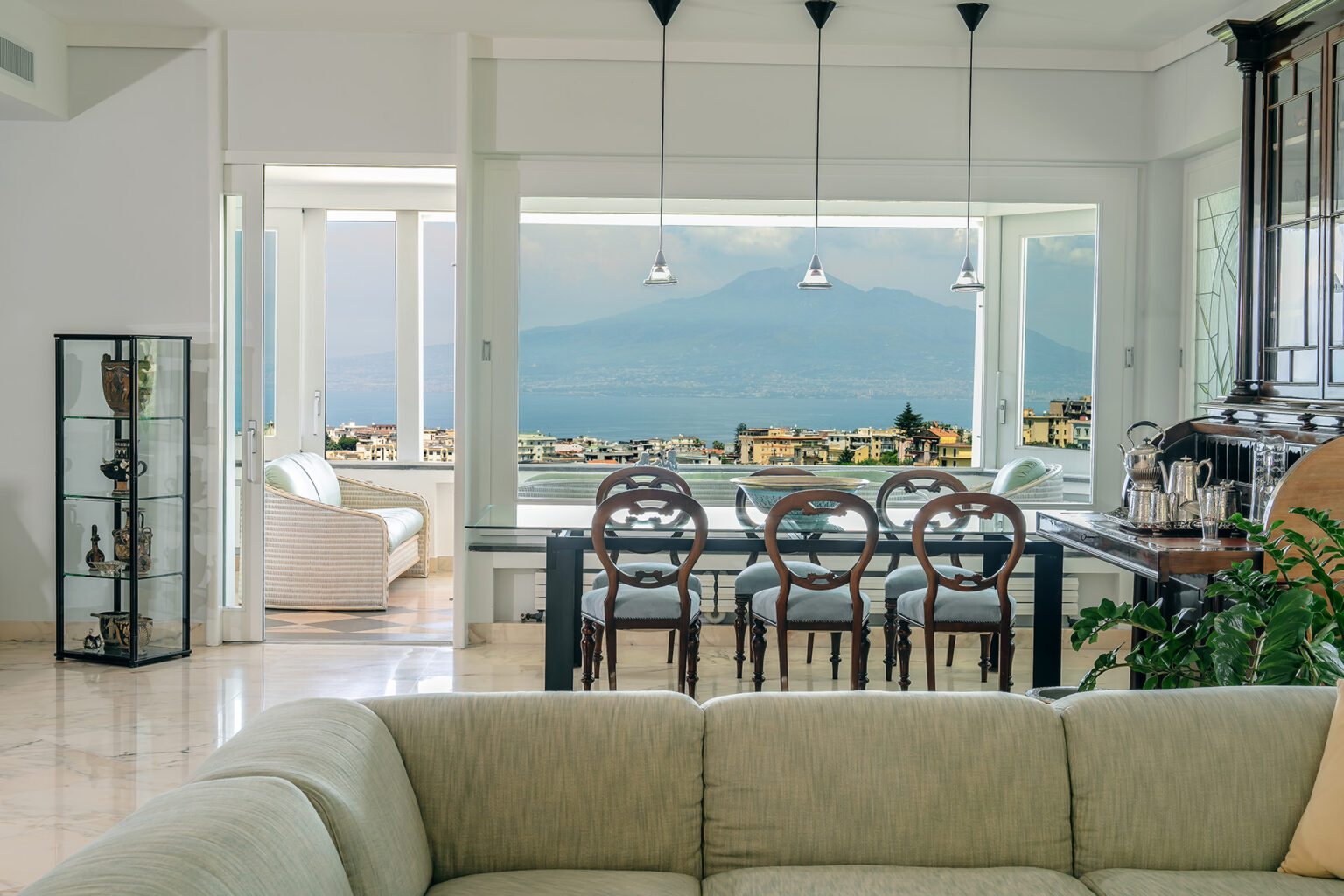 Modern living room with large windows showcasing a view of Mount Vesuvius, featuring an elegant dining area with wooden chairs, a glass table, and decorative items. The space has a light color palette and contemporary furnishings, creating a bright and inviting atmosphere. | Only on SopranoVillas