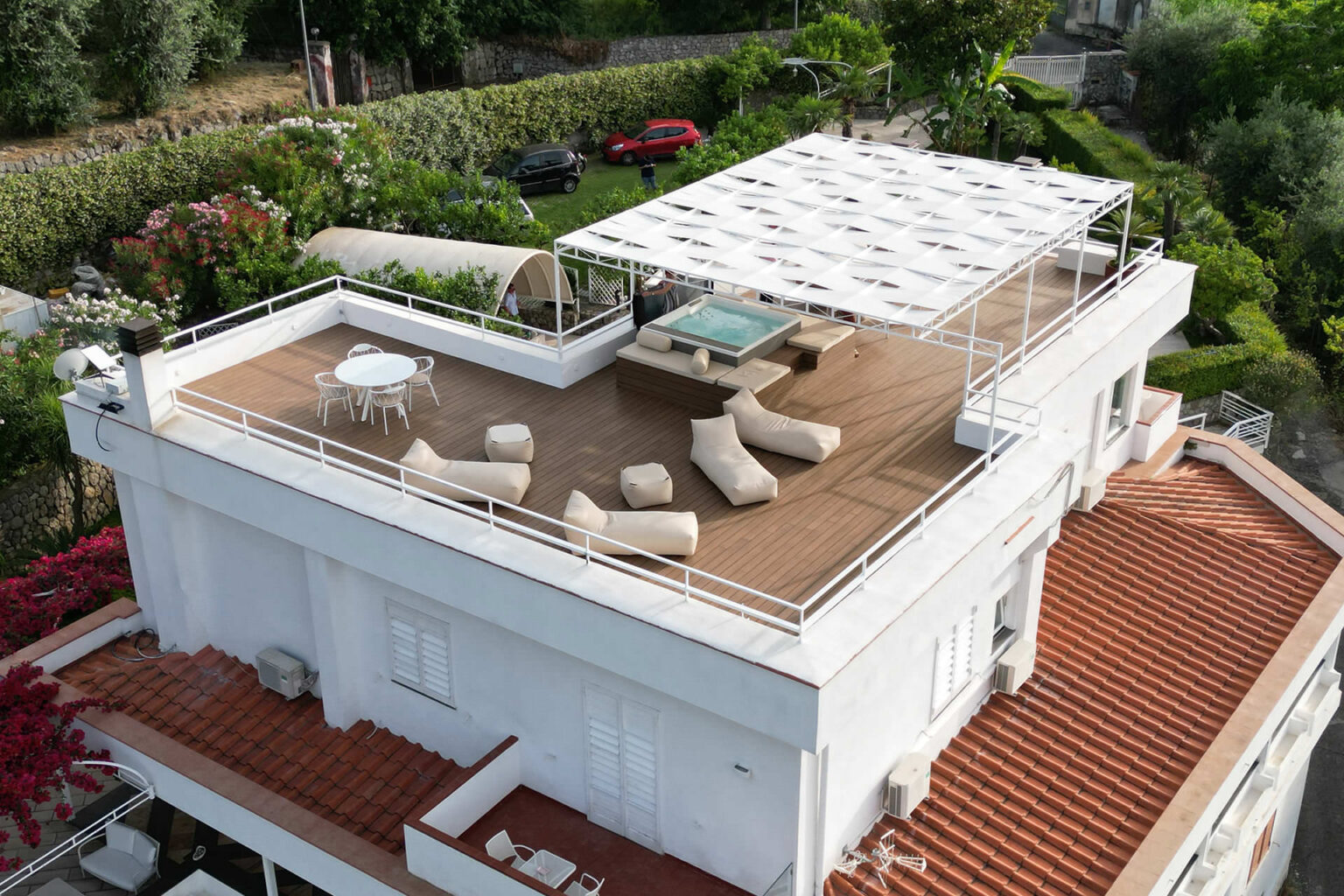 Aerial view of a modern rooftop terrace featuring a shaded seating area with lounge chairs, a small table, and a hot tub surrounded by greenery and flowers. The terrace is designed for relaxation and outdoor enjoyment. | Only on SopranoVillas