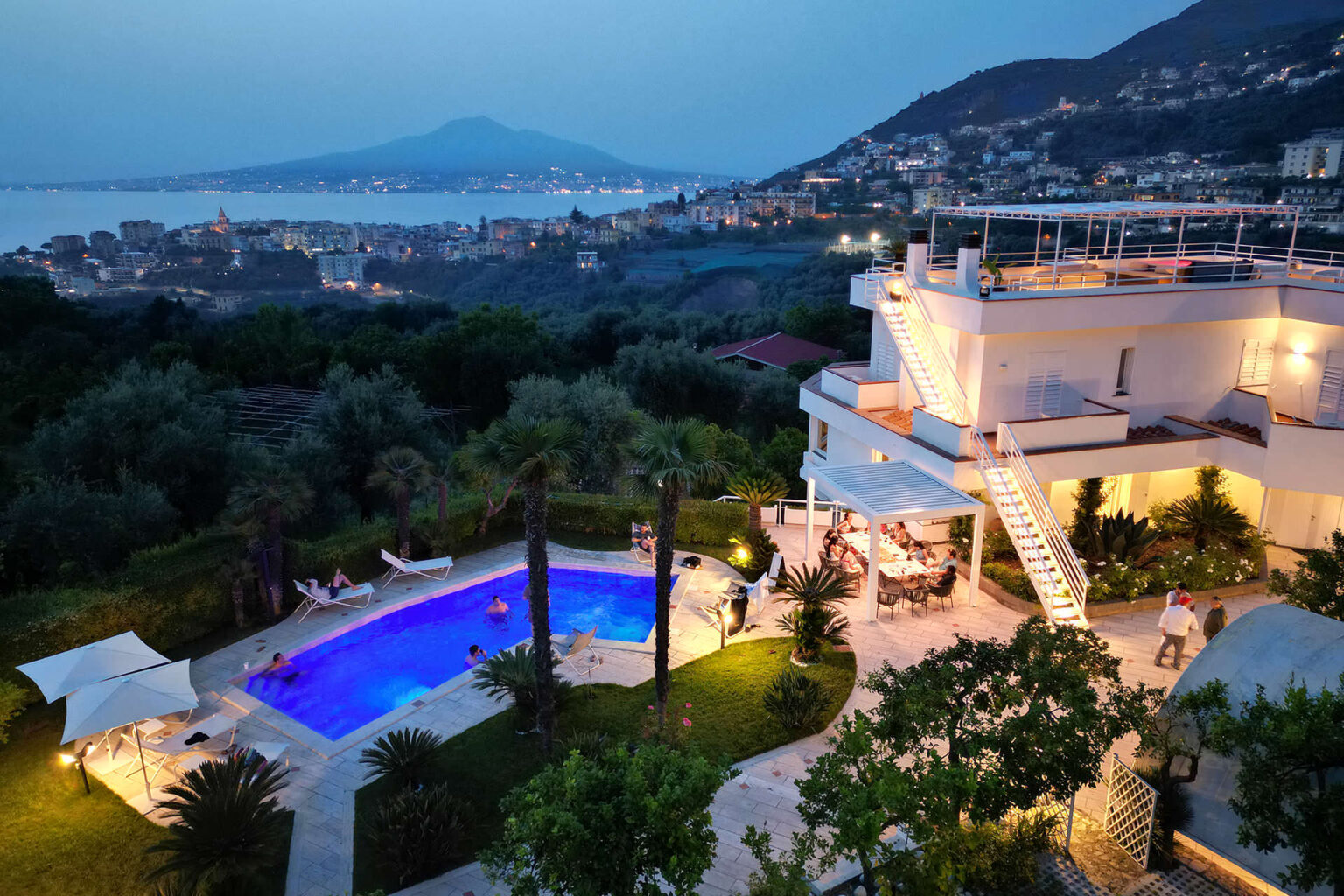 Luxurious villa with a stunning pool and beautiful landscaping, overlooking a coastal city at dusk, with a view of the mountains in the background. | Only on SopranoVillas