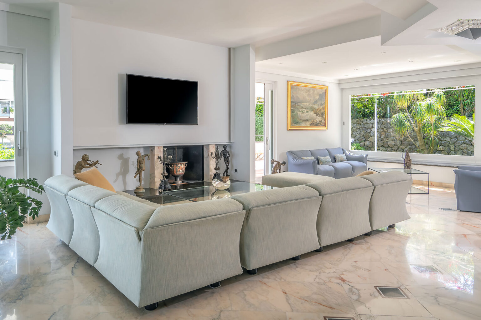 Spacious modern living room featuring a light-colored sectional sofa, a flat-screen TV mounted on the wall, and large windows with views of greenery. Elegant decor includes a decorative fireplace and artwork, creating a bright and inviting atmosphere. | Only on SopranoVillas