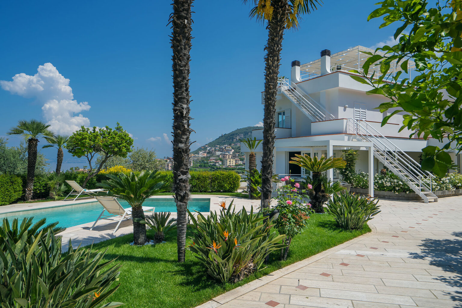Modern villa with a swimming pool surrounded by lush greenery and palm trees, featuring a scenic view of the mountains and clear blue sky. Ideal for luxury living or vacation rentals. | Only on SopranoVillas