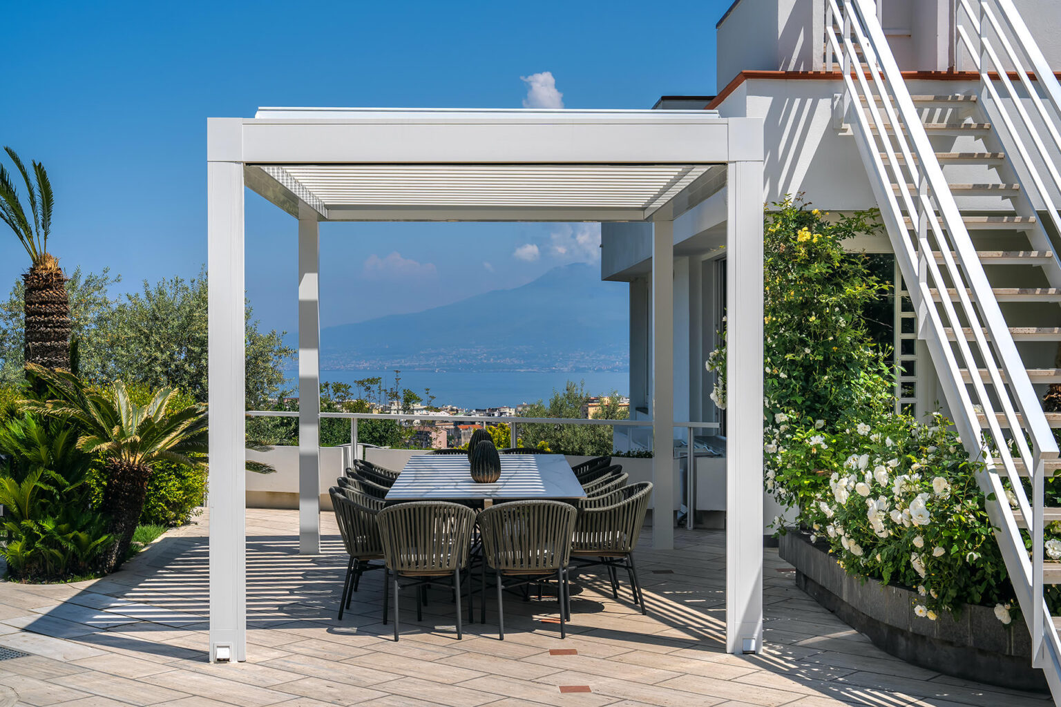 Modern outdoor dining area with a pergola, featuring a large table and chairs, surrounded by lush greenery and a scenic view of the ocean and mountains in the background. | Only on SopranoVillas