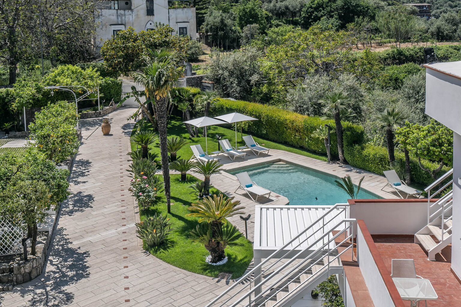 Aerial view of a luxurious villa with a swimming pool surrounded by lush greenery and palm trees, featuring sun loungers under white umbrellas, and a paved pathway leading through a beautifully landscaped garden. Ideal for relaxation and outdoor enjoyment. | Only on SopranoVillas