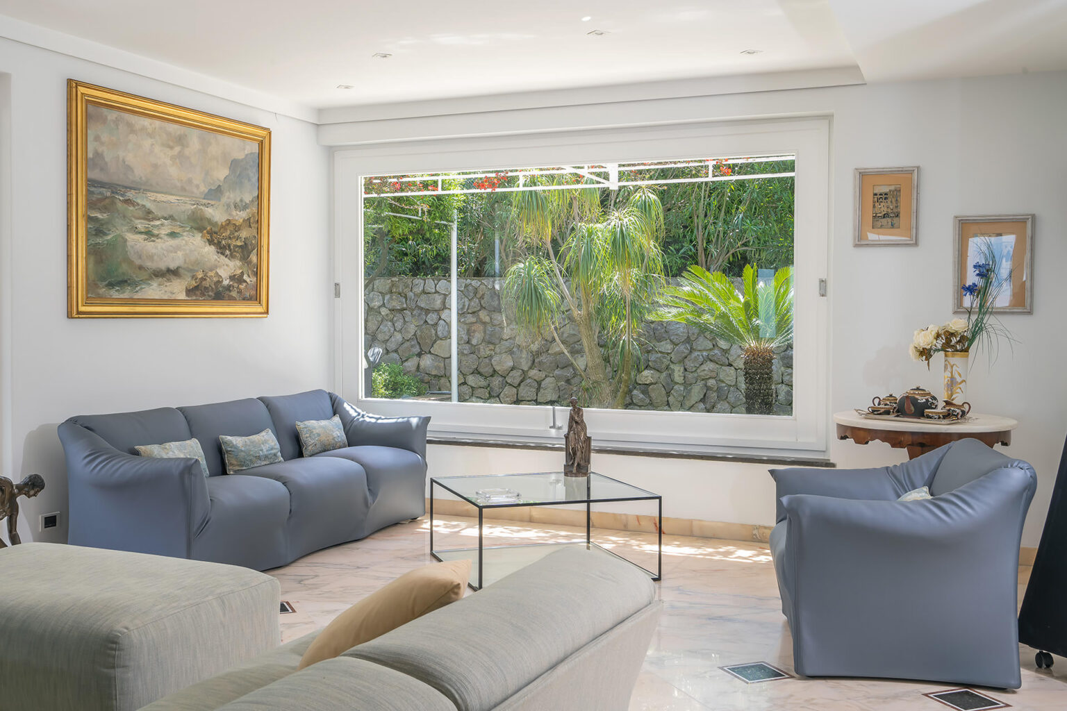 Bright and airy living room featuring a modern design, blue sofas, a glass coffee table, and large windows showcasing lush greenery outside, complemented by decorative art on the walls. | Only on SopranoVillas