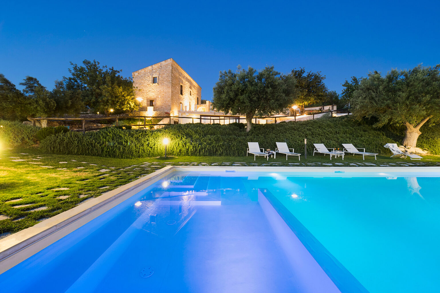 Luxurious villa with a sparkling pool, surrounded by lush greenery and olive trees, illuminated at dusk for a serene getaway experience. | Only on SopranoVillas