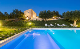 Luxurious villa with a sparkling pool, surrounded by lush greenery and olive trees, illuminated at dusk for a serene getaway experience.