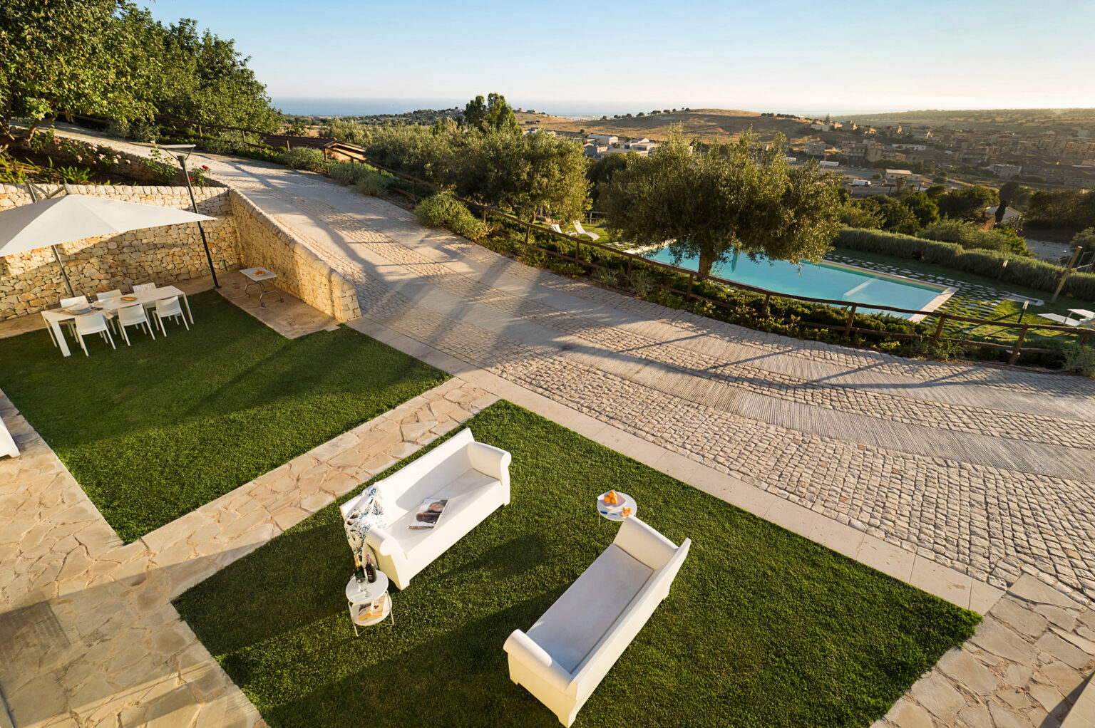 Aerial view of a luxurious outdoor patio featuring modern white sofas and a dining area, surrounded by well-maintained greenery and a swimming pool, with scenic hills in the background under a clear sky. | Only on SopranoVillas
