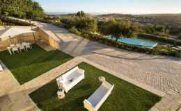 Aerial view of a luxurious outdoor patio featuring modern white sofas and a dining area, surrounded by well-maintained greenery and a swimming pool, with scenic hills in the background under a clear sky.