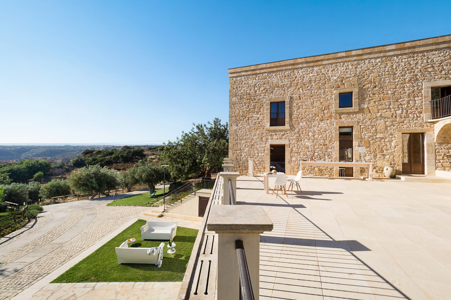 Exterior view of a stone villa with a spacious terrace, overlooking a scenic landscape with olive trees and a gravel path, under a clear blue sky. | Only on SopranoVillas