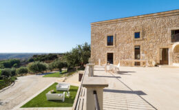Exterior view of a stone villa with a spacious terrace, overlooking a scenic landscape with olive trees and a gravel path, under a clear blue sky.