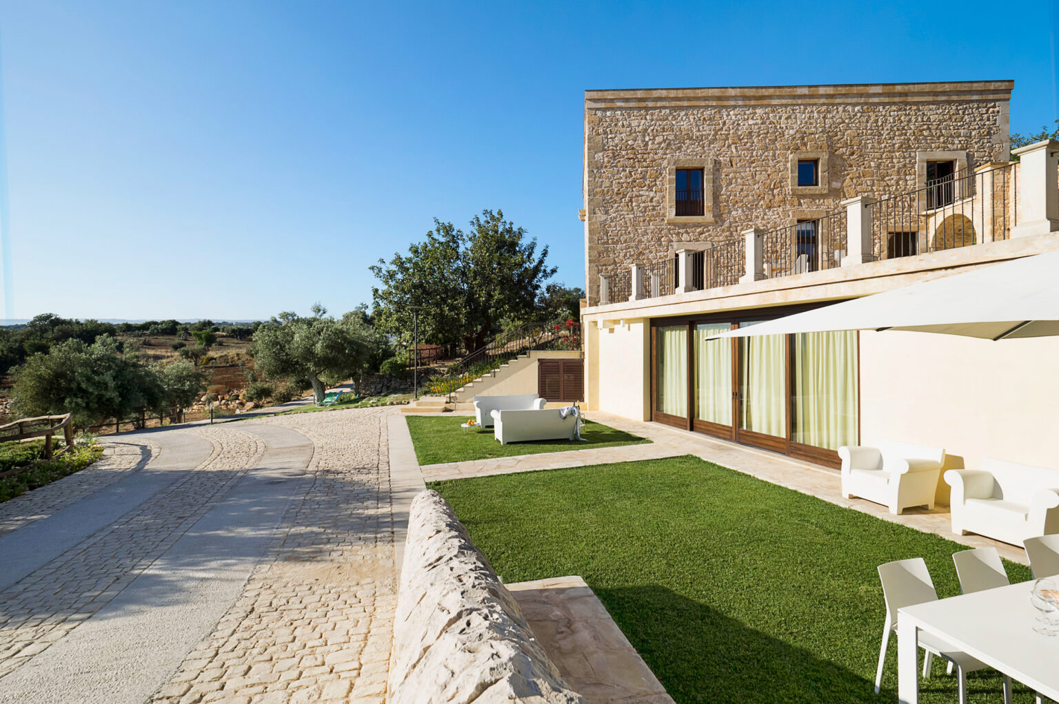 A scenic view of a modern stone building with large windows, surrounded by lush greenery and a well-maintained lawn. The image features a clear blue sky and a pathway leading up to the structure, highlighting the tranquil outdoor setting. Ideal for showcasing architectural design and nature integration. | Only on SopranoVillas