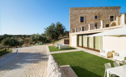A scenic view of a modern stone building with large windows, surrounded by lush greenery and a well-maintained lawn. The image features a clear blue sky and a pathway leading up to the structure, highlighting the tranquil outdoor setting. Ideal for showcasing architectural design and nature integration.