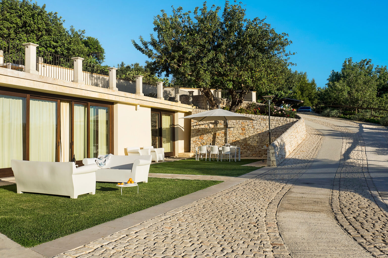 Modern villa exterior with a landscaped garden, outdoor seating area, and a winding stone pathway leading up a hillside. Lush greenery surrounds the property, providing a serene and inviting atmosphere. | Only on SopranoVillas