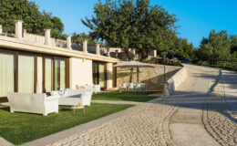 Modern villa exterior with a landscaped garden, outdoor seating area, and a winding stone pathway leading up a hillside. Lush greenery surrounds the property, providing a serene and inviting atmosphere.