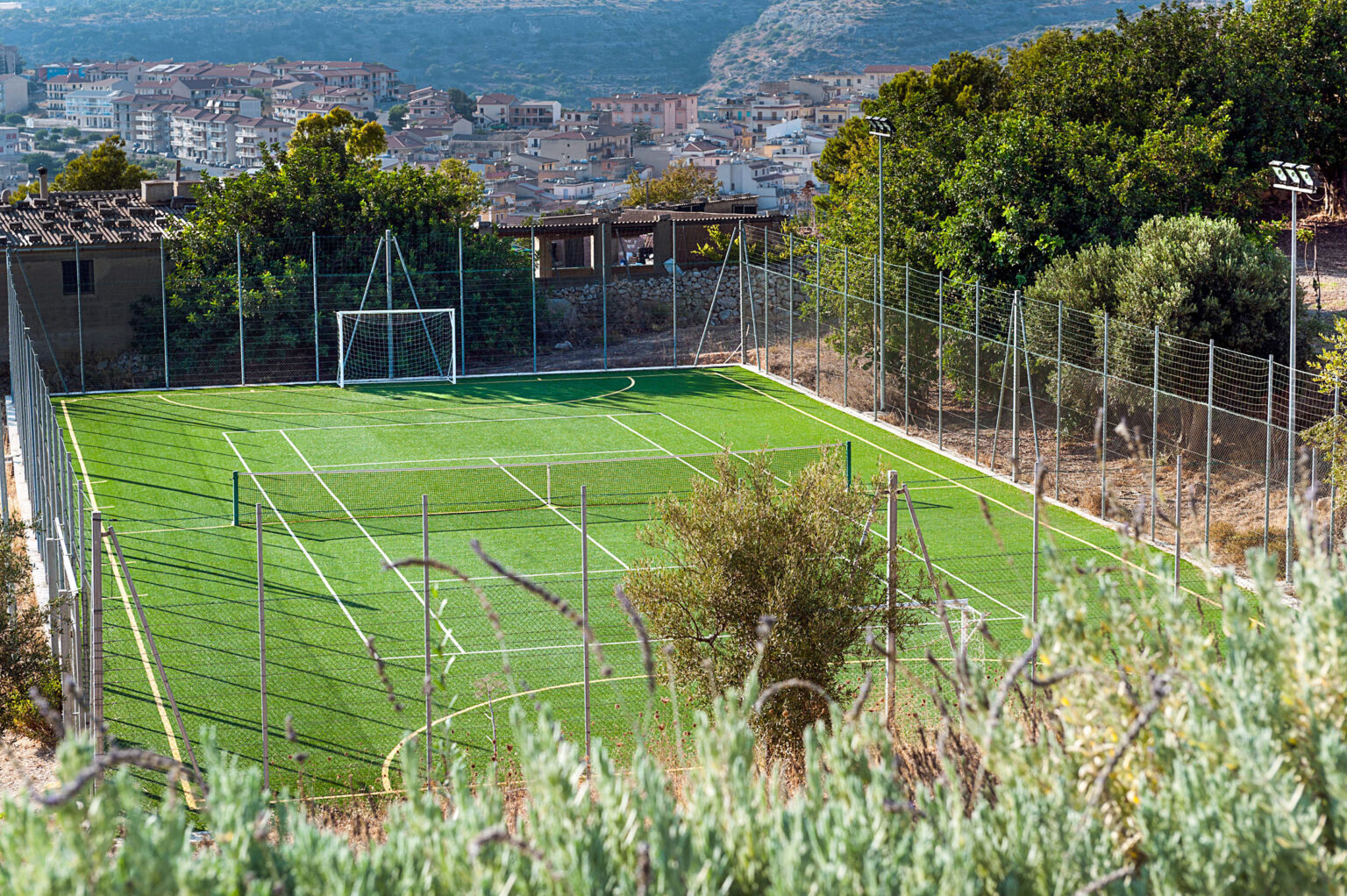 Aerial view of a small, well-maintained soccer field surrounded by a fence, set against a backdrop of a city and mountains. The artificial turf is bright green, and goalposts are visible at either end of the field. | Only on SopranoVillas