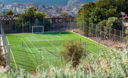 Aerial view of a small, well-maintained soccer field surrounded by a fence, set against a backdrop of a city and mountains. The artificial turf is bright green, and goalposts are visible at either end of the field.