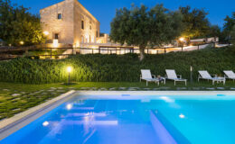 Scenic view of a luxurious stone villa at dusk, featuring an inviting swimming pool and comfortable lounge chairs surrounded by lush greenery and illuminated by soft outdoor lighting.